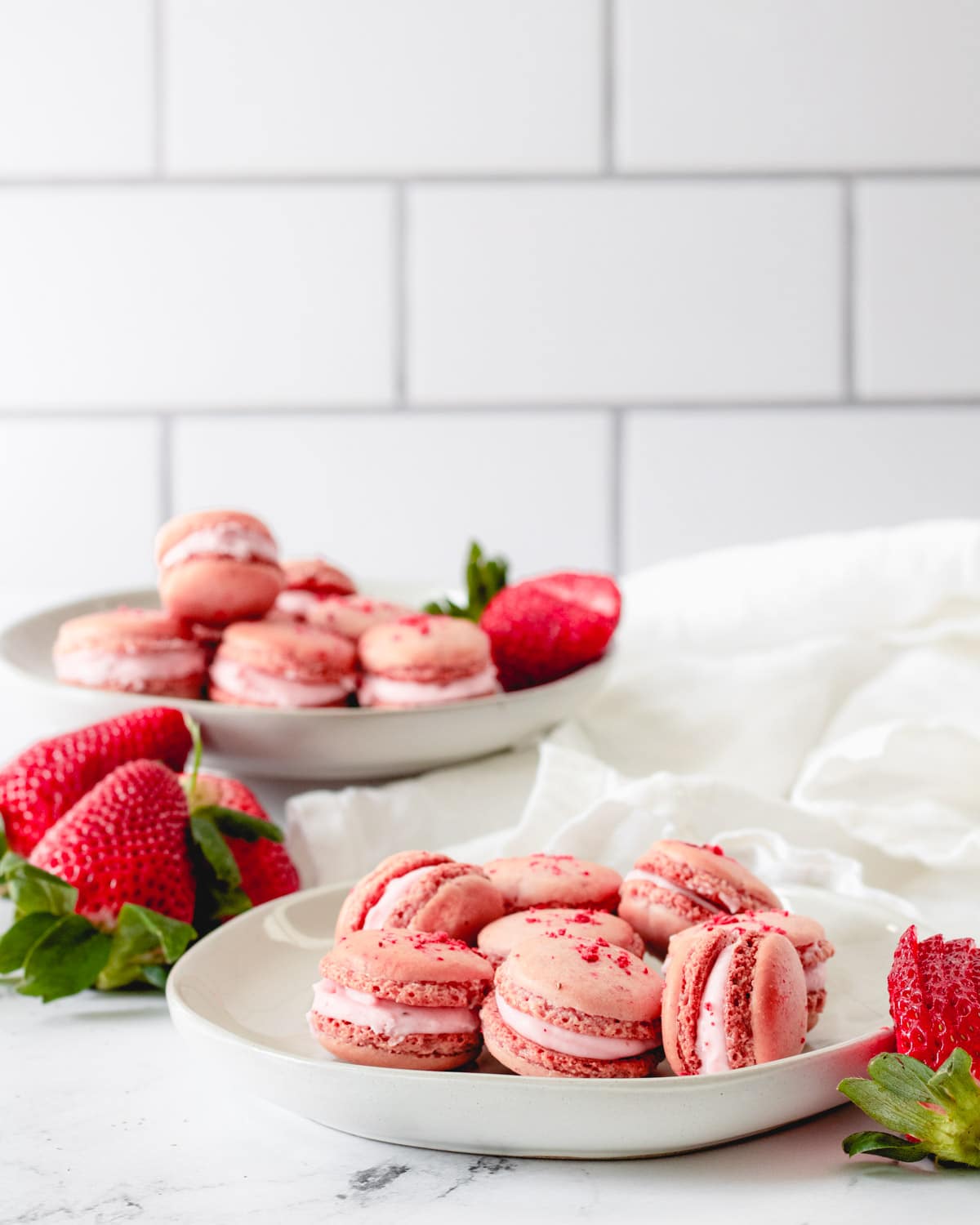 Two plates of strawberry macarons with some fresh strawberries scattered around.