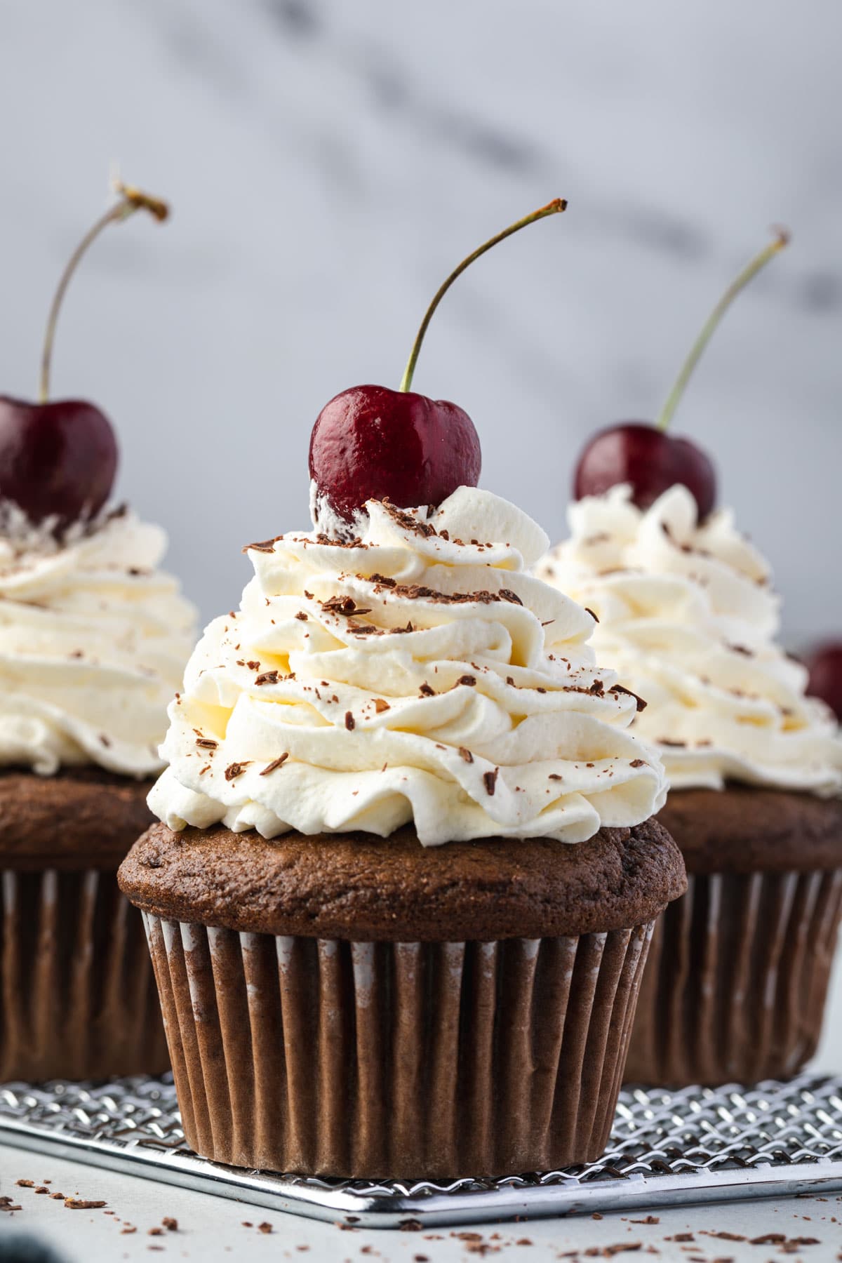 3 black forest cupcakes on cooling rack topped with whipped cream & cherry.