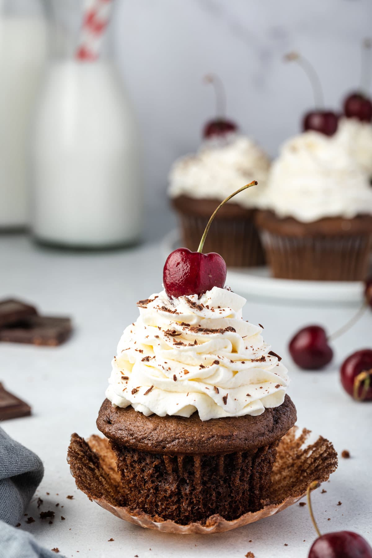 Black forest cupcake with liner peeled off and a plate of cupcakes & jar of milk in background.