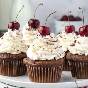 5 Black forest cupcakes on a serving plate with bowl of cherries and milk jug in background.
