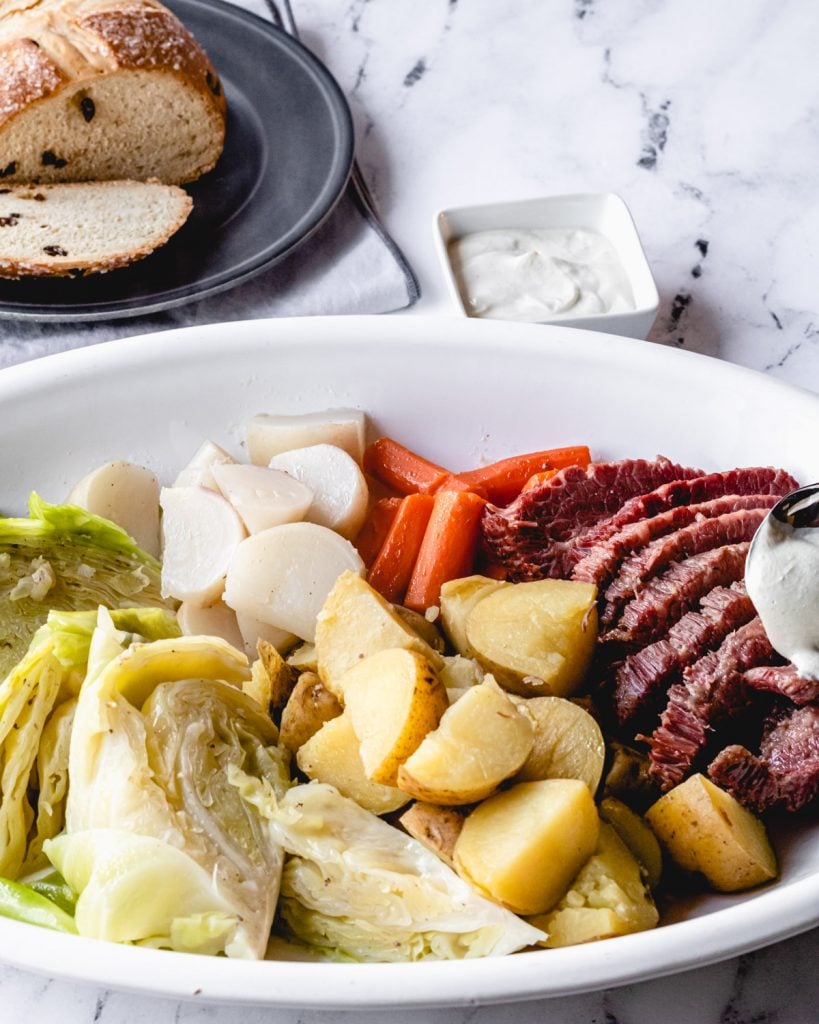 Platter of corned beef with vegetables, and behind it is a ramekin of mustard cream sauce and Irish soda bread on a plate.