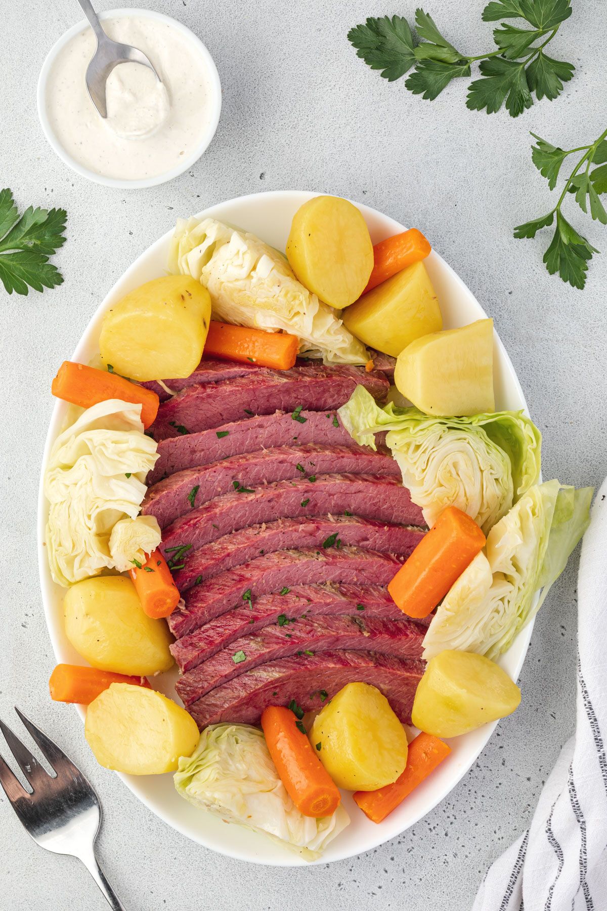 Overhead of a platter of corned beef with veggies surrounding it, parsley scattered, and a bowl of creamy white mustard sauce.