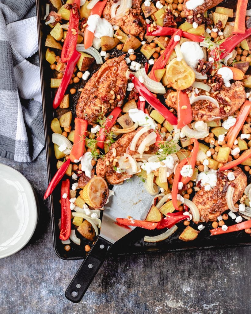Sheet pan with Greek chicken, peppers, potatoes, onions, and a greek yogurt dressing, spatula and a stack of plates.