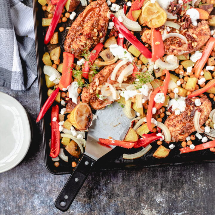 Sheet pan with Greek chicken, peppers, potatoes, onions, and a greek yogurt dressing, spatula and a stack of plates.