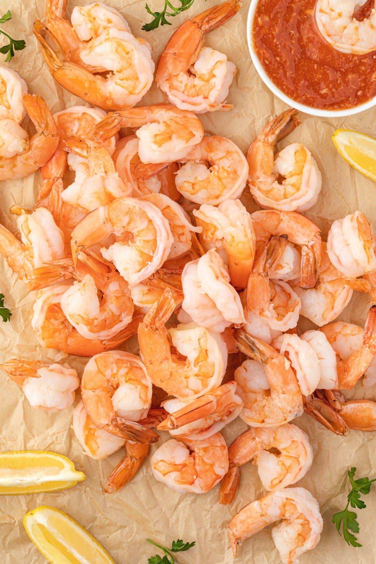 A pile of blanched shrimp on brown parchment with a bowl of cocktail sauce and some lemon wedges.