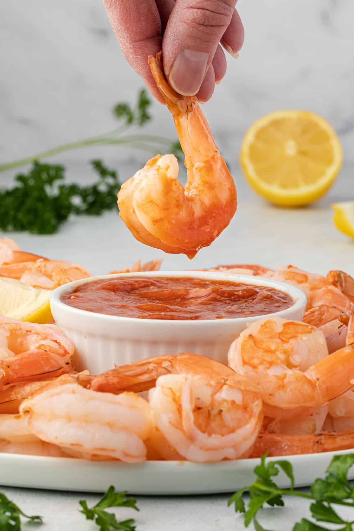 Dipping poached shrimp into a bowl of cocktail sauce that is on a platter filled with shrimp.