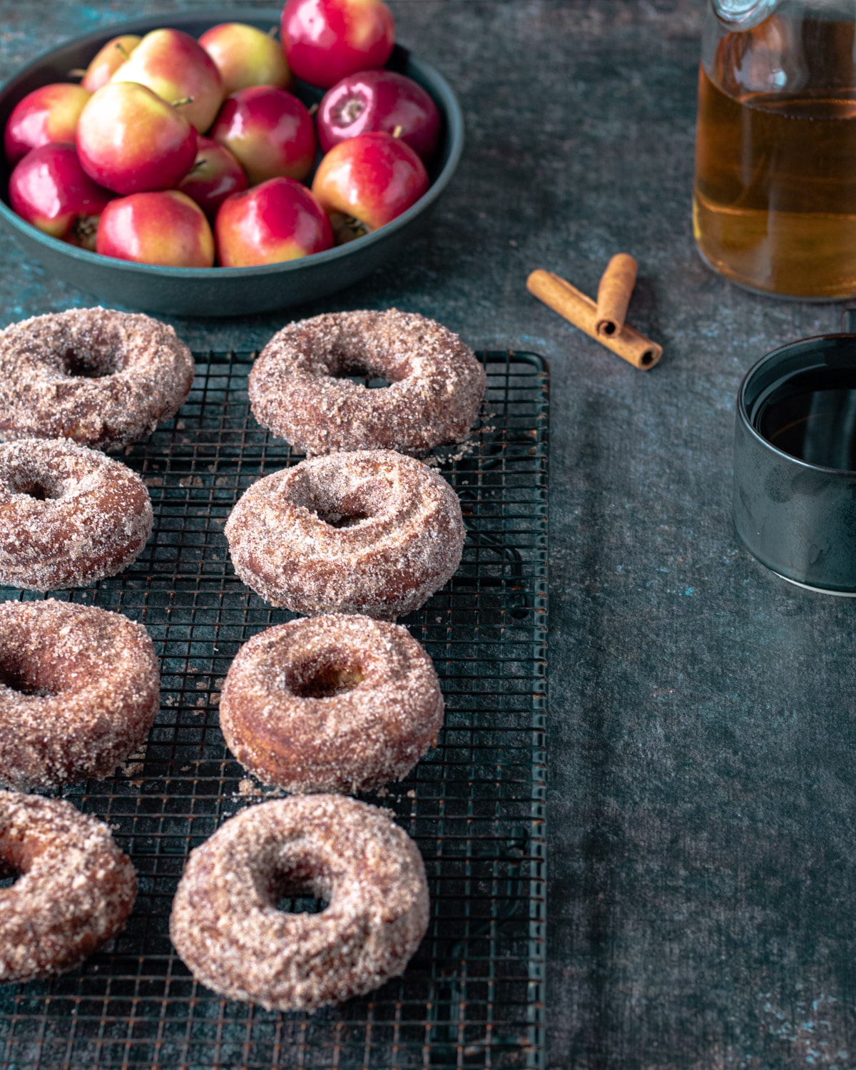 Two rows of apple cider donuts, with a bowl of apples, cinnamon sticks, and cider.