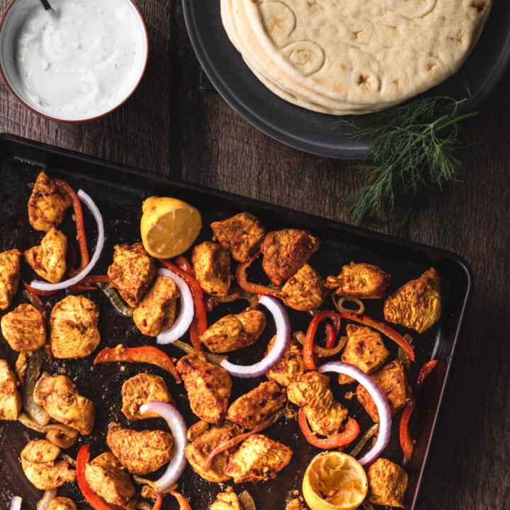 Sheetpan of chicken schwarma with a plate of pitas and a bowl of dill yogurt sauce.