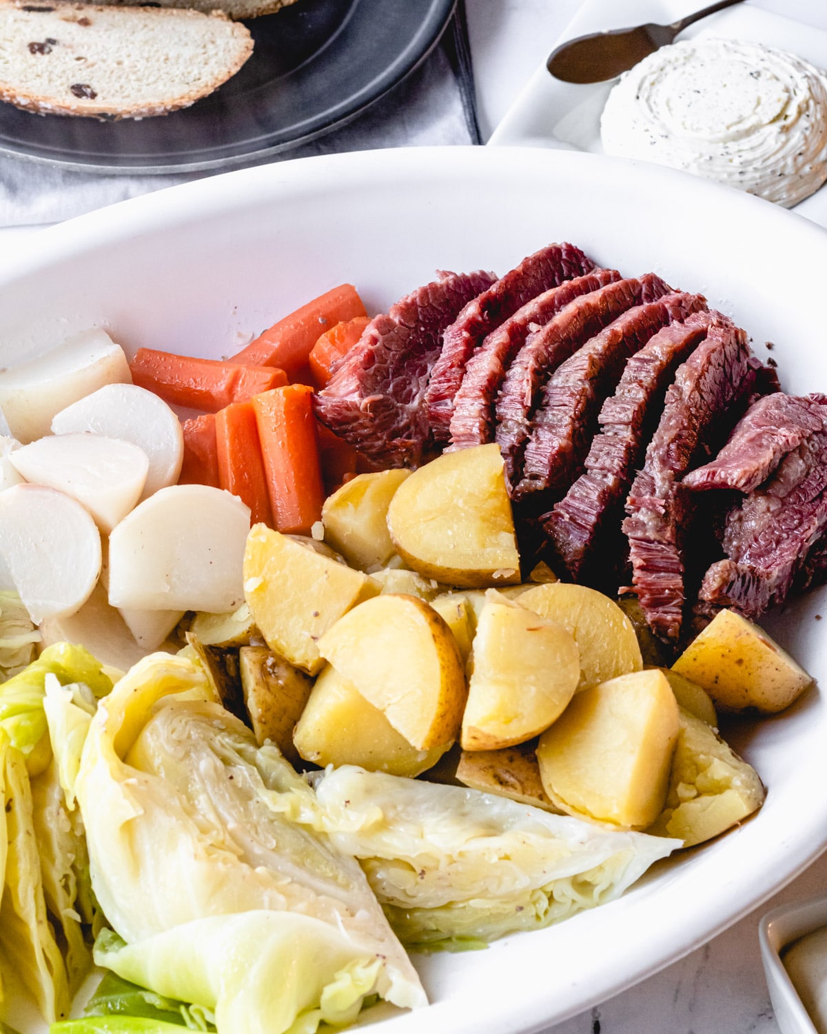 Corned beef on a platter with boiled cabbage, carrots, potatoes, and turnips, with butter and Irish soda bread in background.