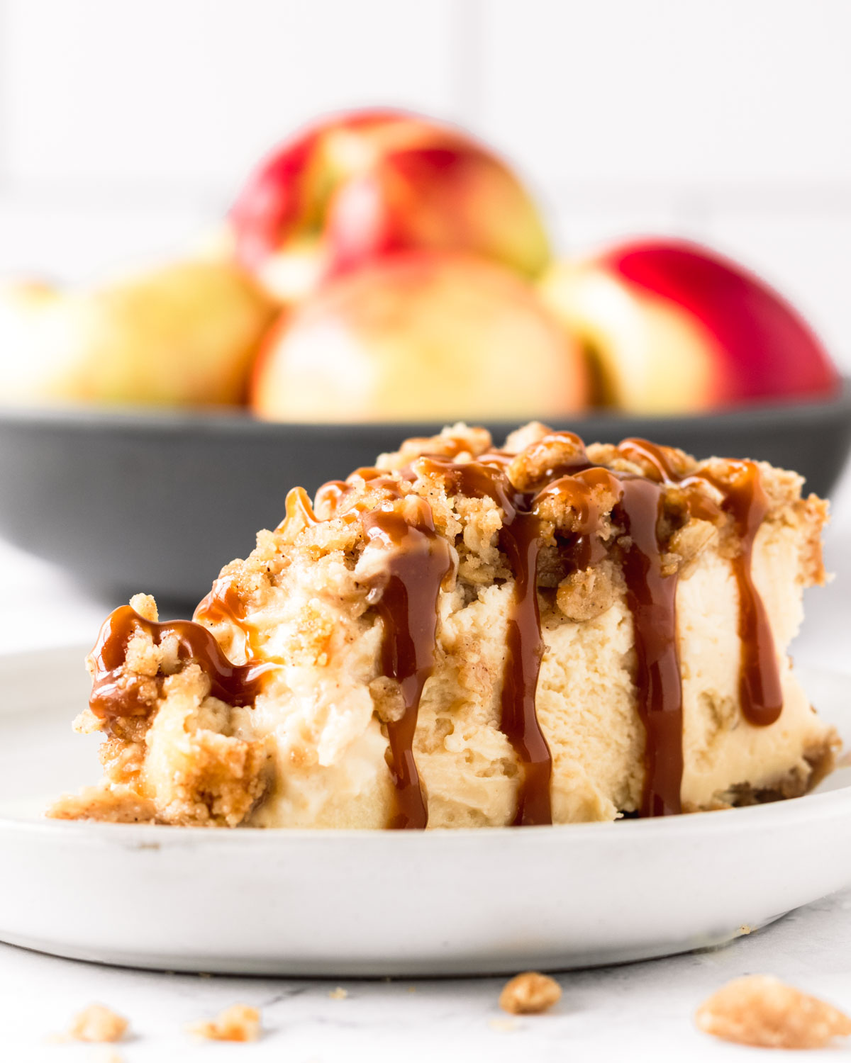 Slice of caramel apple crumble cheesecake with a bowl of apples in the background.