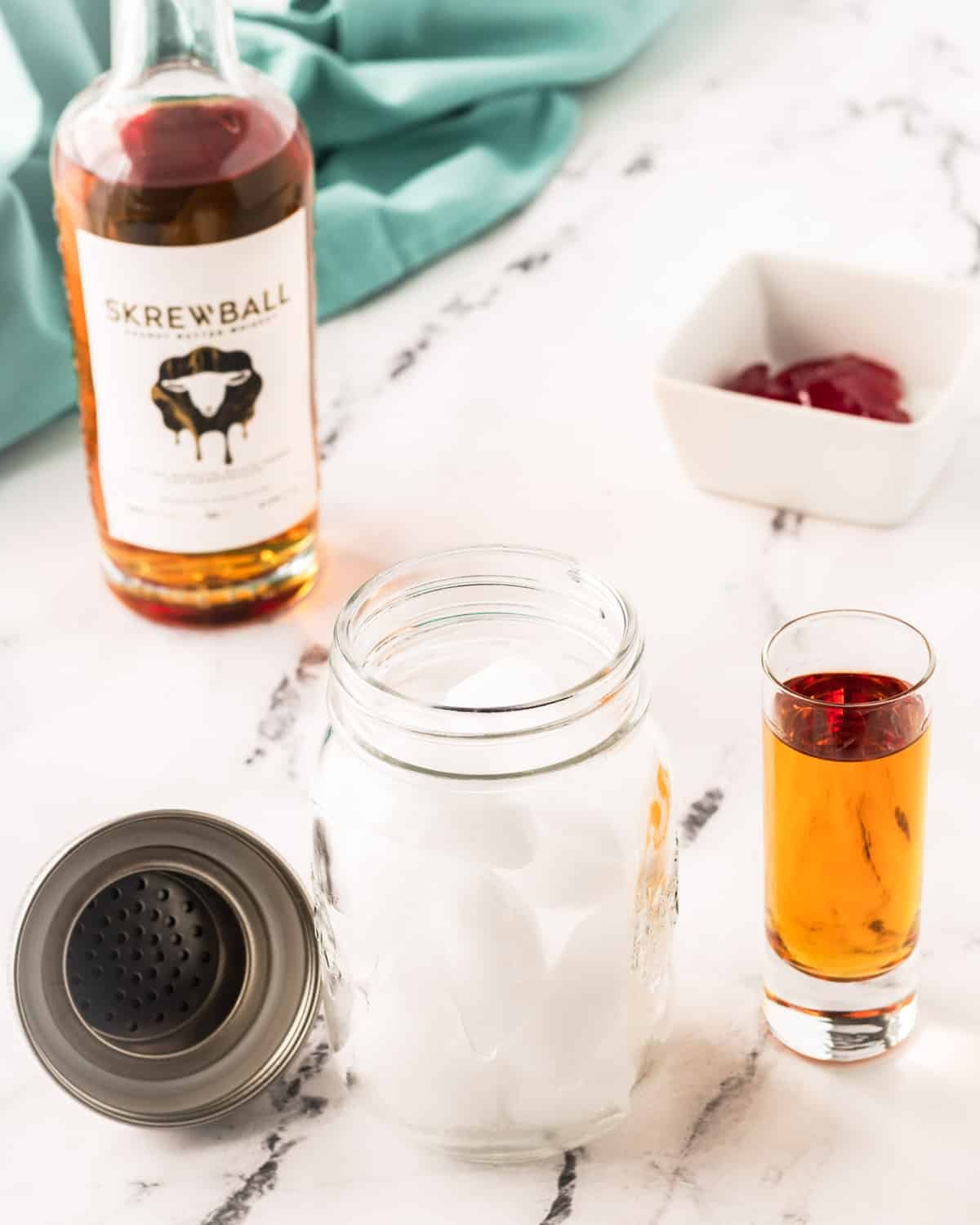 Ice in a mason jar shaker, with bottle of  Skrewball whiskey, strawberry jelly, and a shot glass of whiskey.