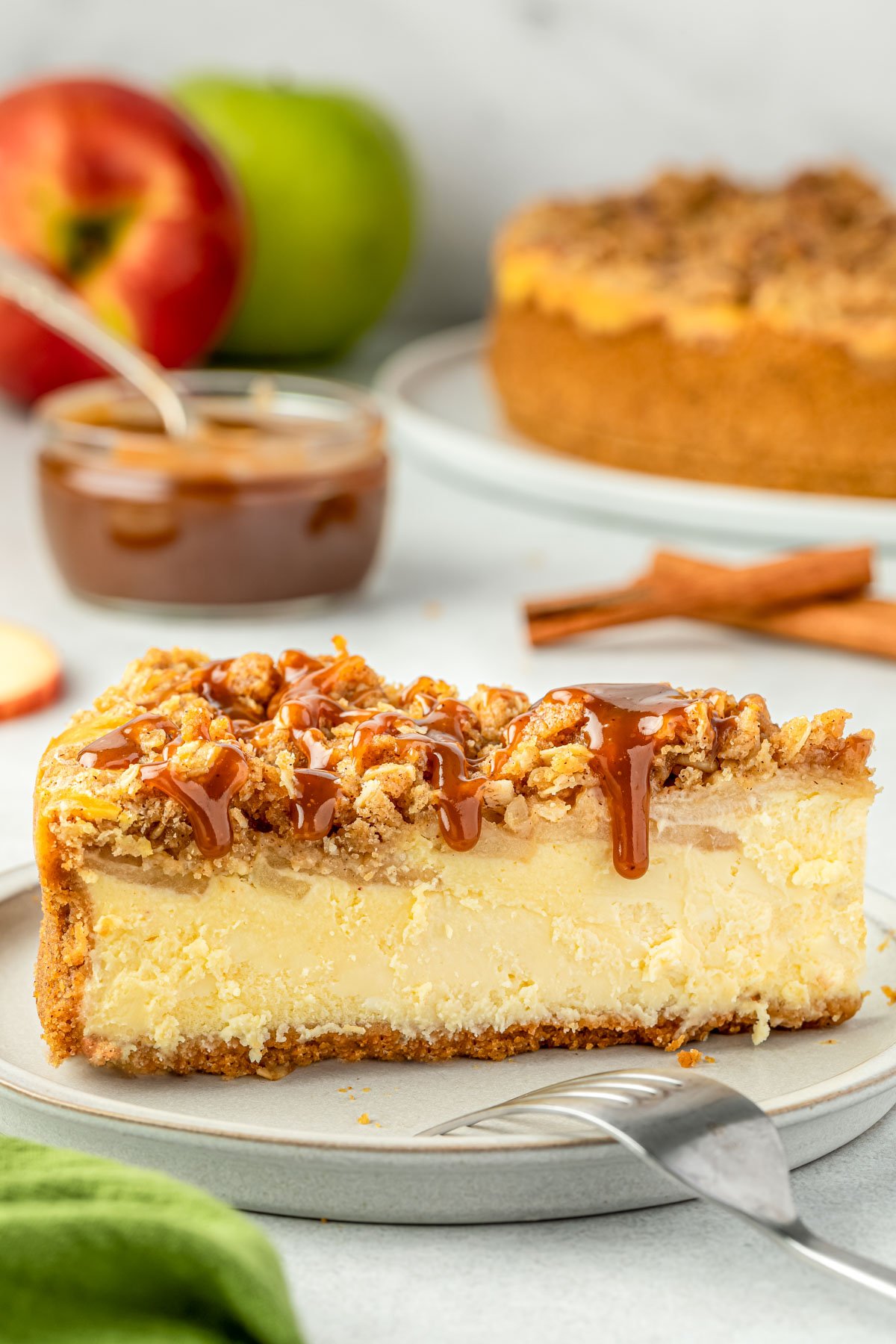 Slice of apple crumble cheesecake with a bowl of caramel sauce, a few apples, and the rest of the cheesecake in the background.