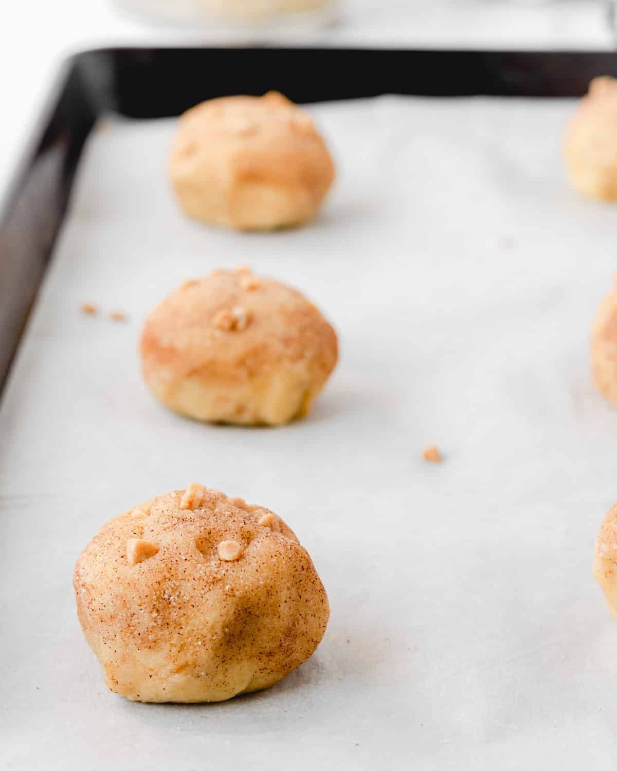 Dough rolled in cinnamon sugar with toffee bits on top, in a row on a cookie sheet.