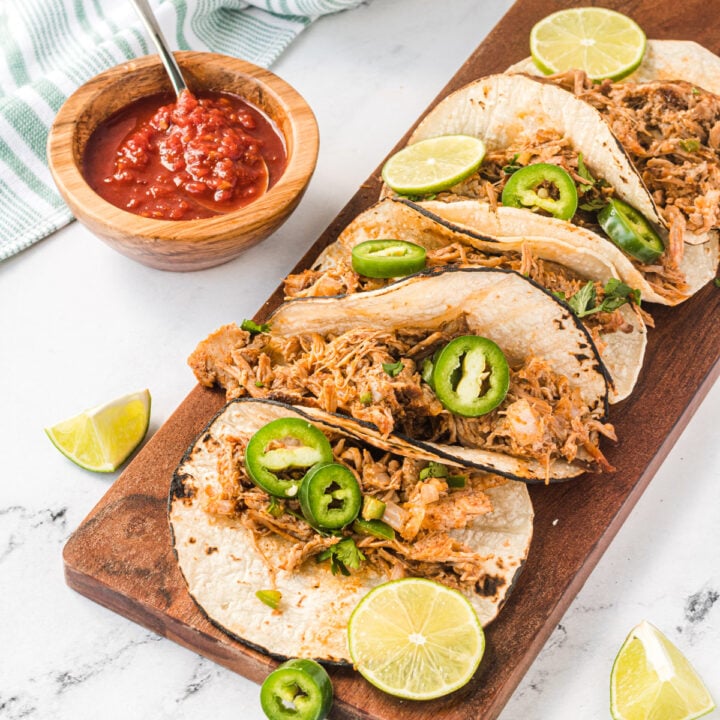 5 carnitas tacos on a wooden platter, garnished with jalapenos and limes.