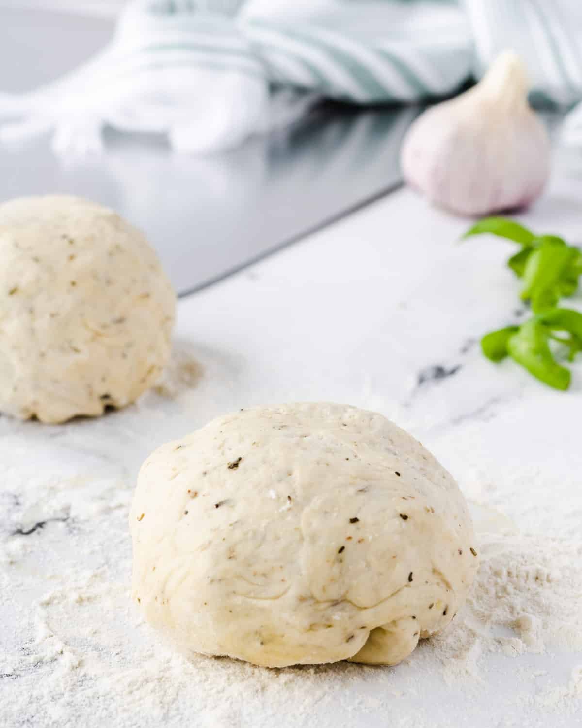 Two balls of garlic herb pizza dough with a bulb or garlic and a sprig of basil in the background.