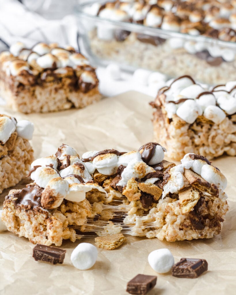 Squares of s'mores rice krispie treats on brown parchment, and the square in the front is split apart showing gooey marshmallow.