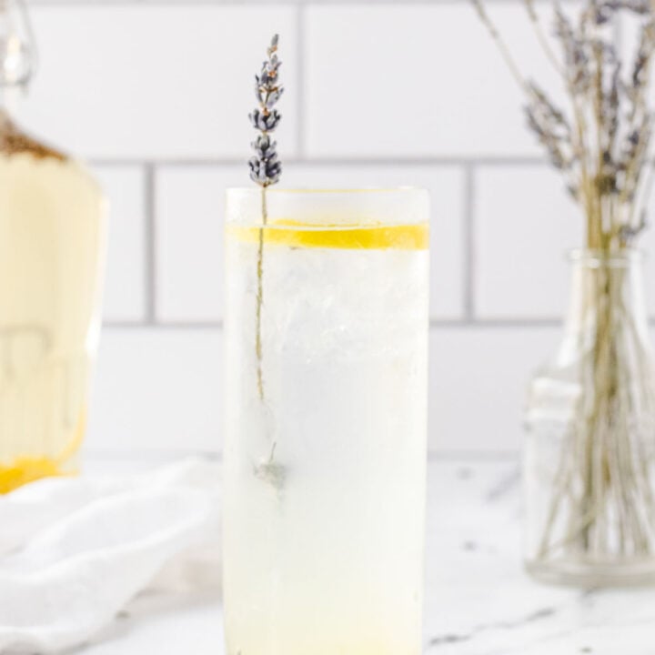 Highball glass of lavender vodka collins, with jar of lavender vodka and vase of lavender stems in background.