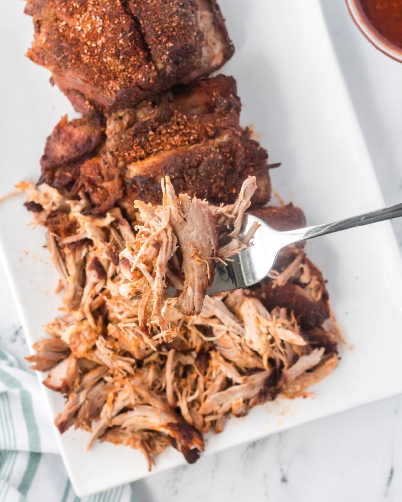 Cooked pork shoulder on a white rectangular plate, with a bite of pork on a fork.