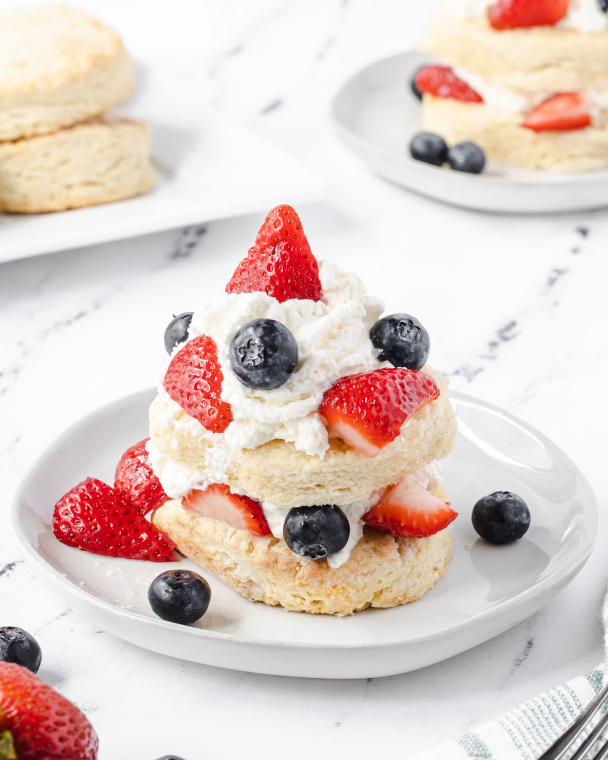 Shortcakes layered with whipped cream, strawberries, and blueberries. A tray of shortcakes and second plate of blueberry strawberry shortcake is in the background.