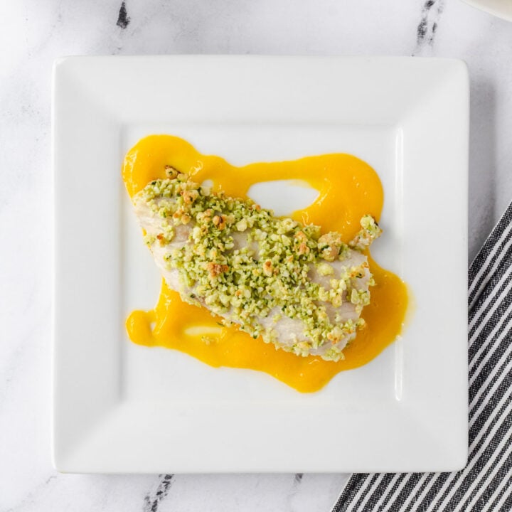 Coconut crusted mango is sitting on mango coulis on a white square plate.