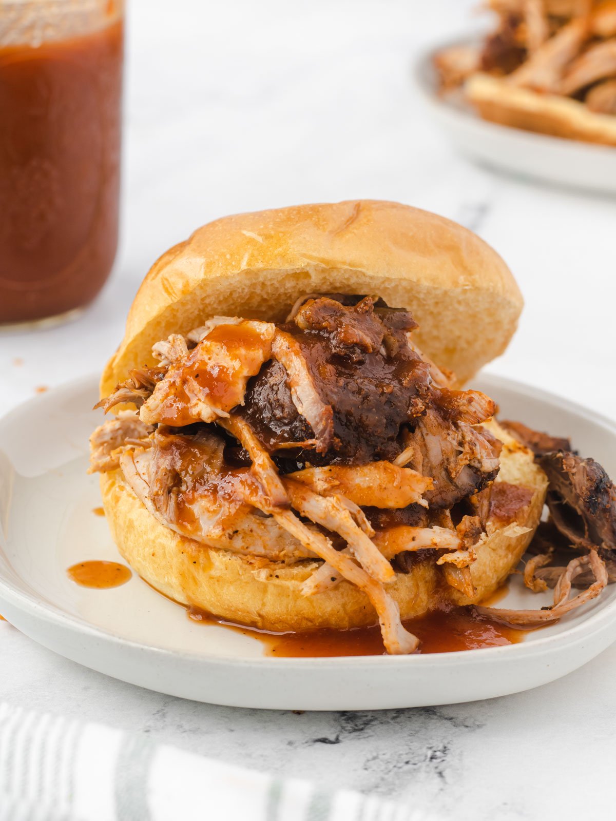 Pulled pork sandwich with pools of bbq sauce and a jar of sauce in the background.