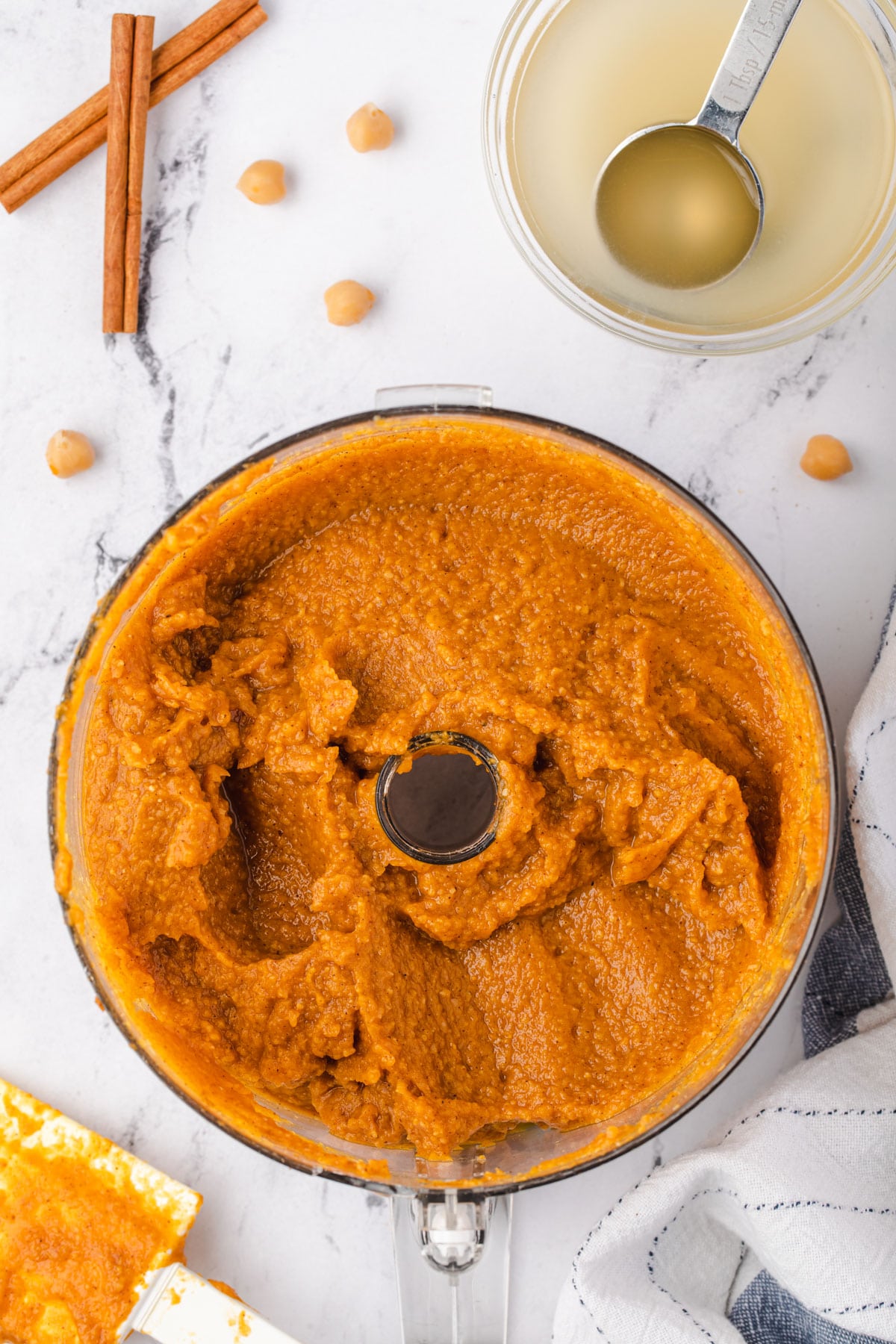 Pumpkin hummus in food processor with bowl of chickpea liquid in corner.
