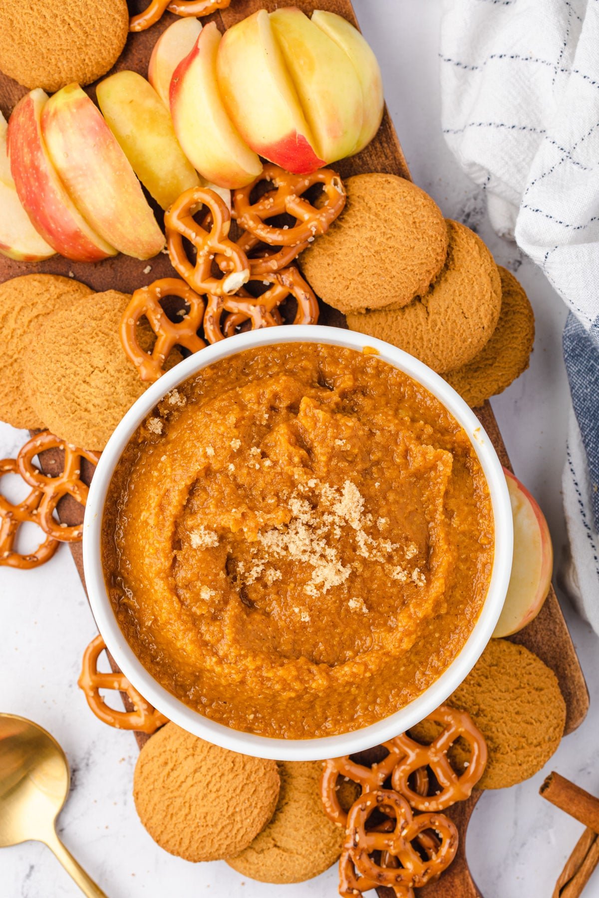 Overhead of bowl of pumpkin hummus sitting on a wooden board with gingersnaps, apples, and pretzels on the board.