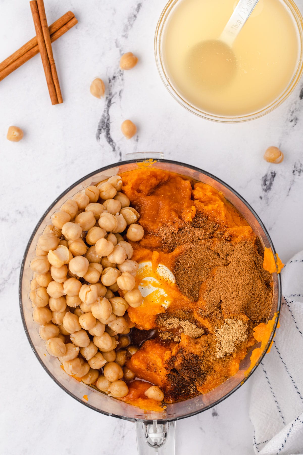 Hummus ingredients in food processor with reserved chickpea liquid in a bowl.