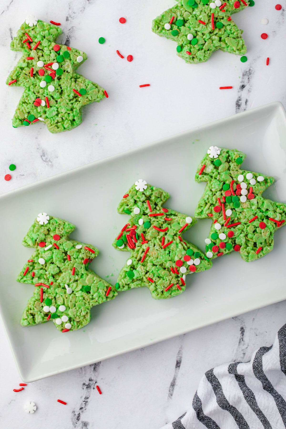 3 Christmas Tree rice krispie treats on a rectangle plate with sprinkles around.
