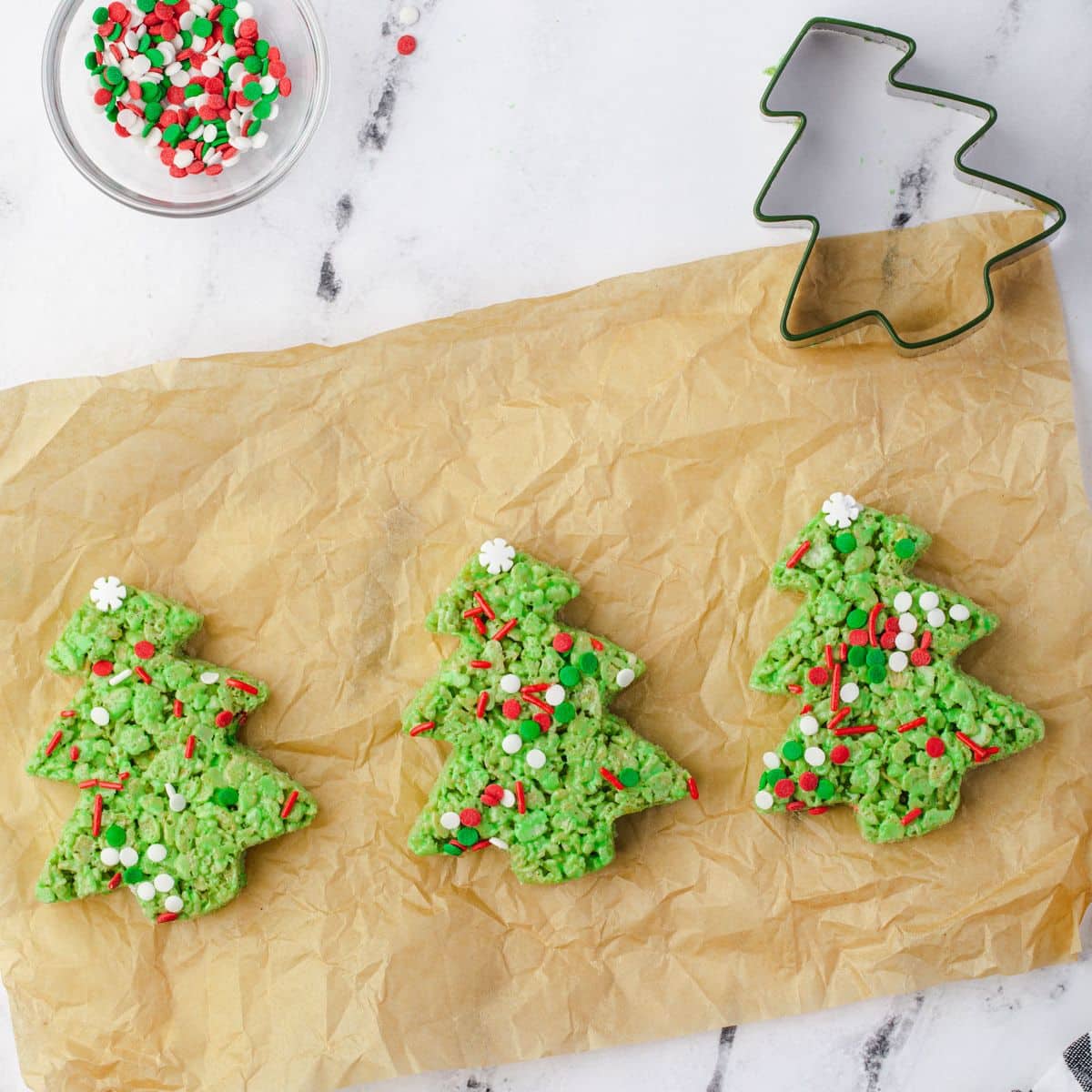 Three decorated Christmas tree Rice Krispie treats on parchment paper with sprinkles and a tree-shaped cookie cutter.