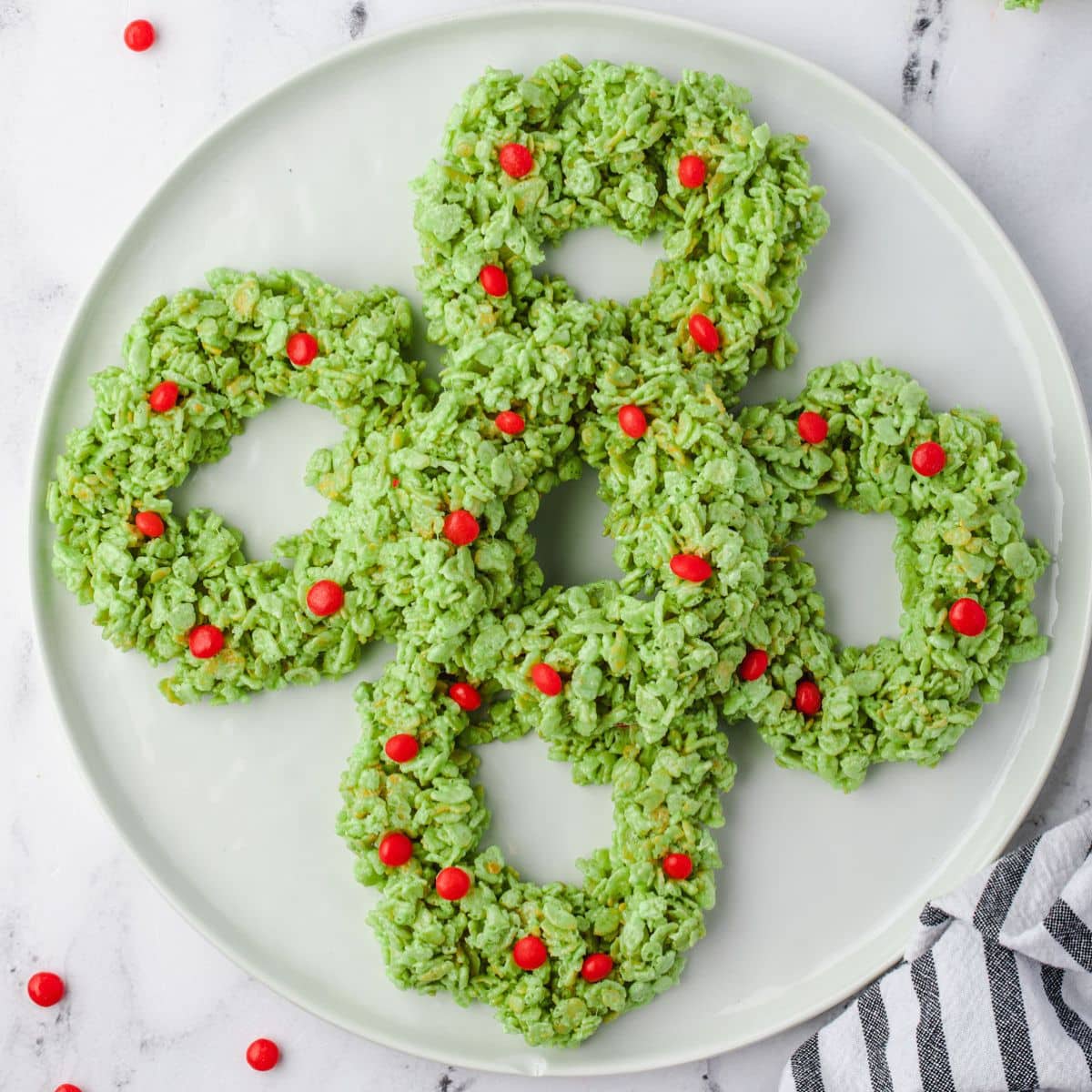 Green rice krispie wreaths with red candies on a white plate, arranged in a clover shape.