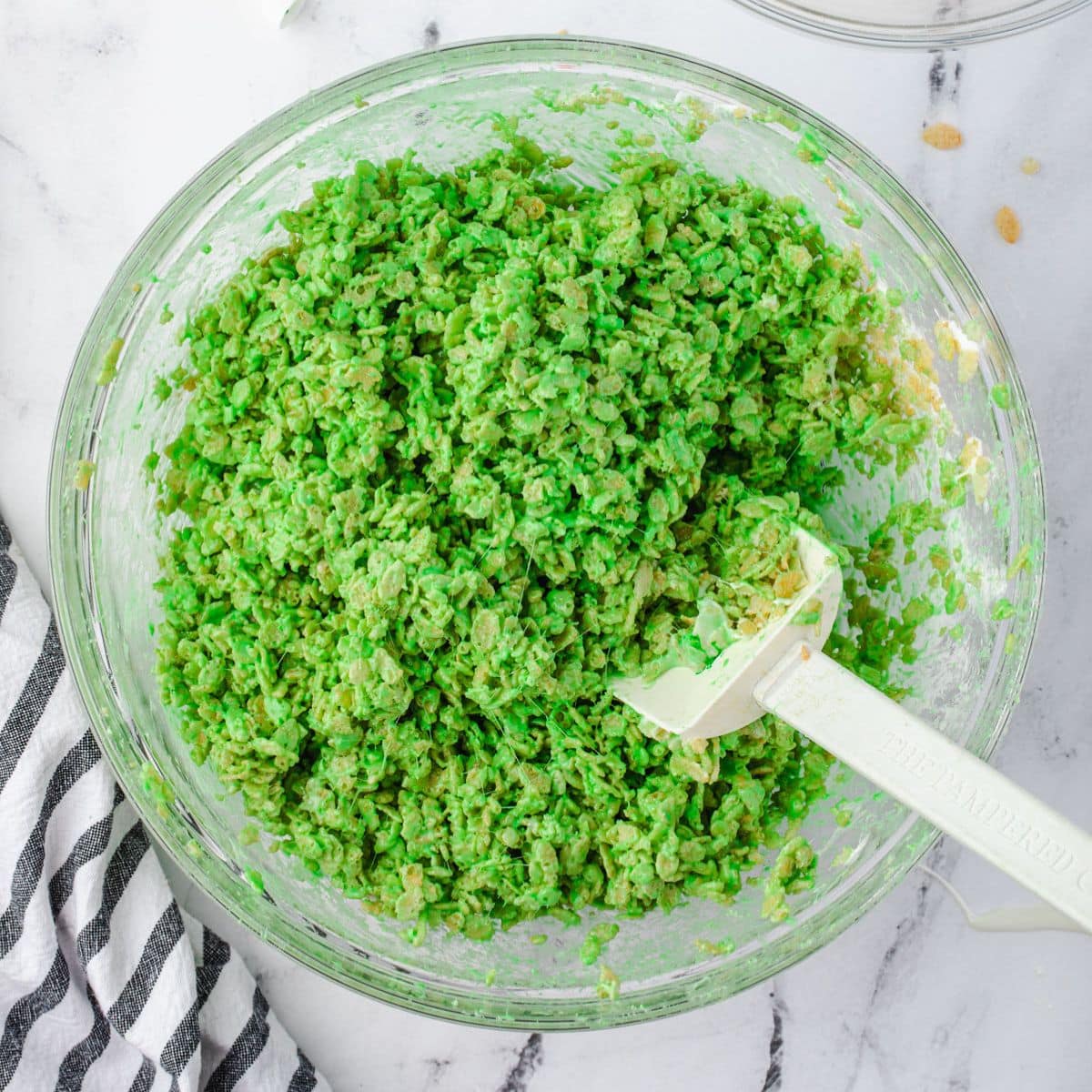 A glass bowl of bright green marshmallow coated cereal with a spatula inside, on a marble countertop.