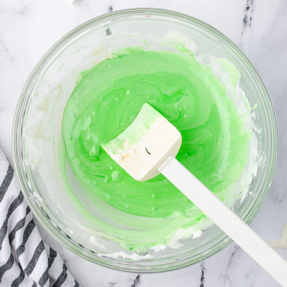 A bowl of green melted marshmallows with a white spatula on a marble surface.