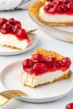 No-bake cheesecake on a plate, topped with cherry pie filling, and another slice behind it.