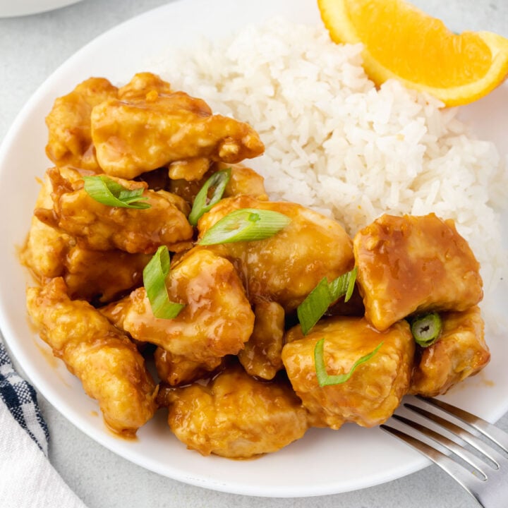 Plate or orange chicken topped with green onions, rice, orange wedge, and fork.
