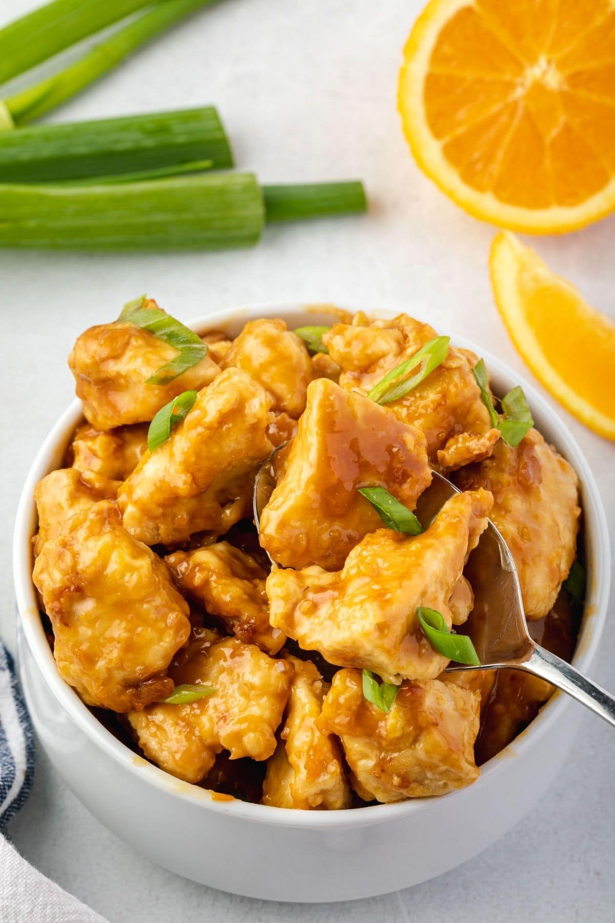 Bowl of orange chicken with piece on fork, green onions and orange half on top.