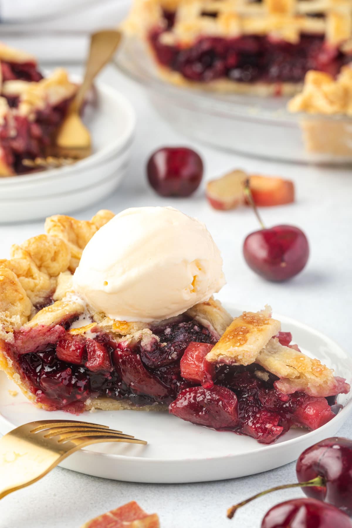 Slice of rhubarb cherry pie on a plate with a scoop of ice cream and whole pie in background.