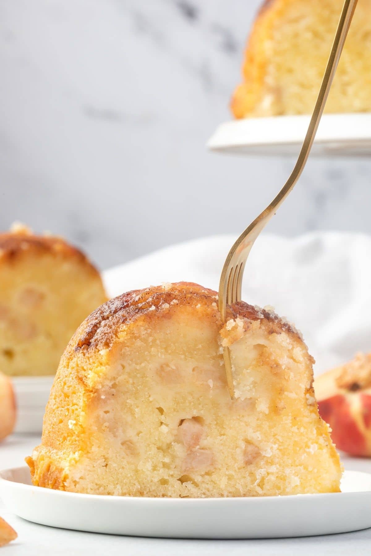 Slice of pound cake with chopped peaches in it, standing upright on a plate and a gold fork through the top portion taking a bite.