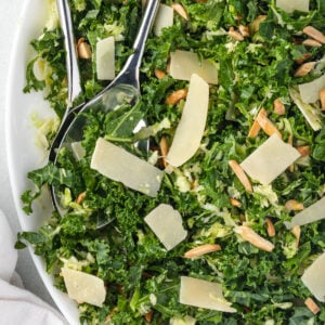 Close up of a kale salad with stainless serving tools, topped with shaved parmesan and almonds.