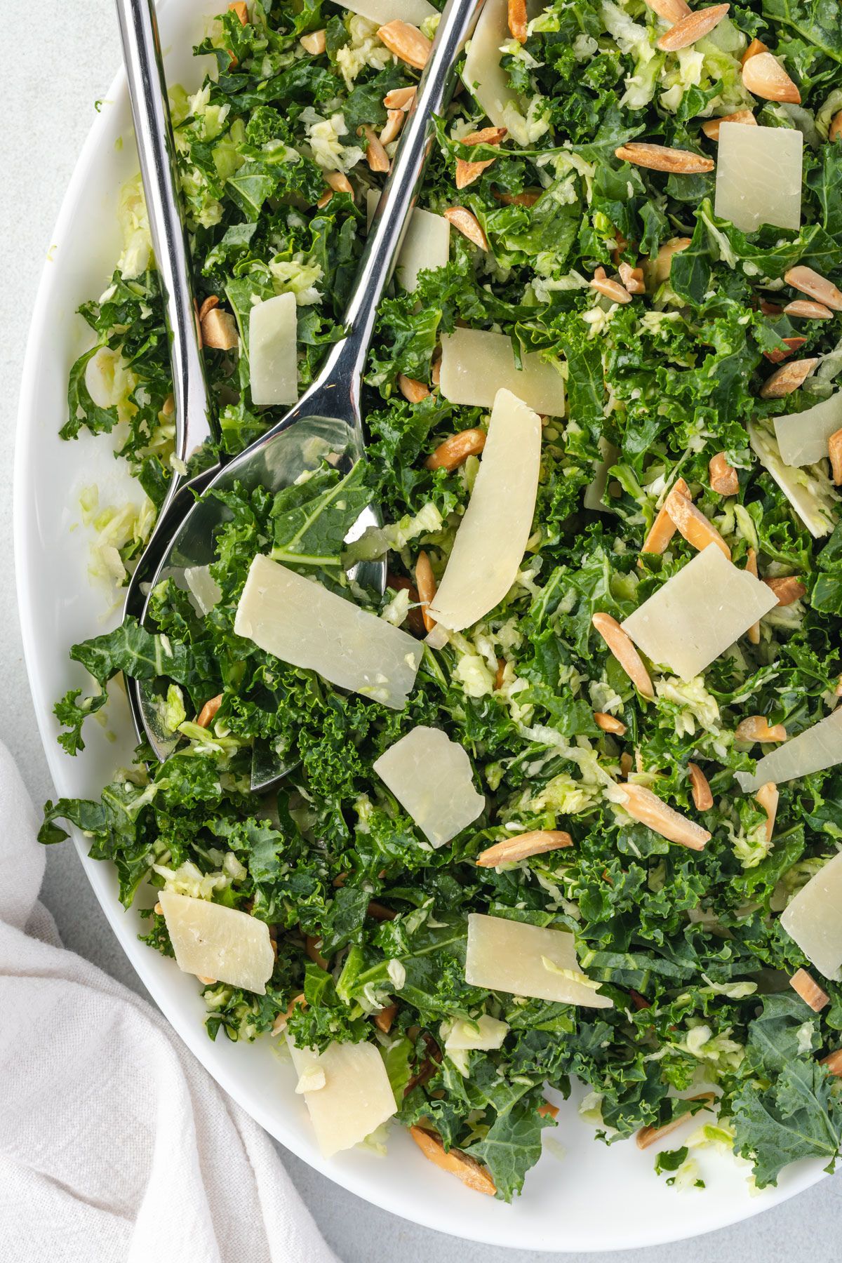 Close up of a kale salad with stainless serving tools, topped with shaved parmesan and almonds.