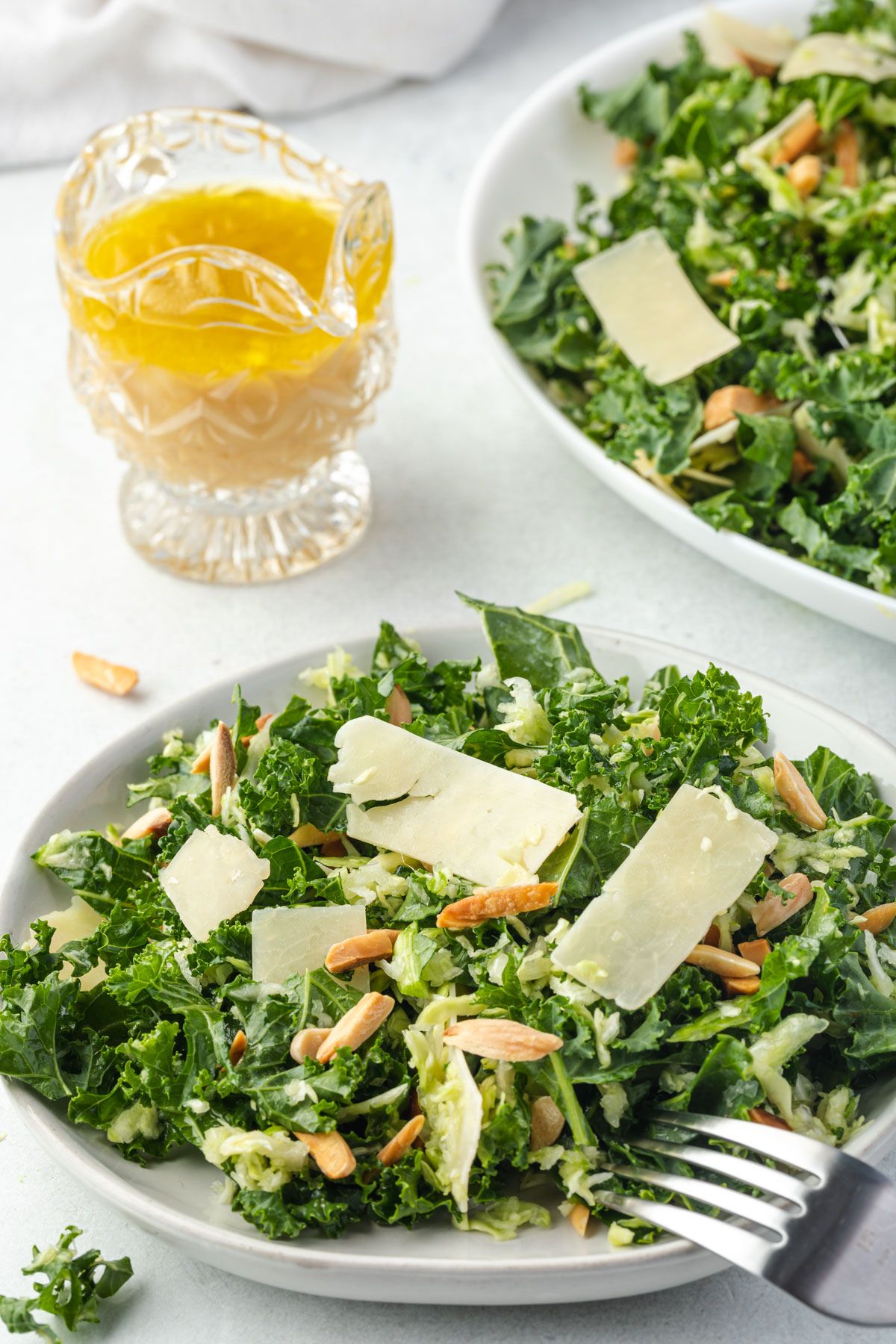 Kale salad with parmesan & almonds on a plate, with a fork resting on side, and crystal pitcher of dressing in back.