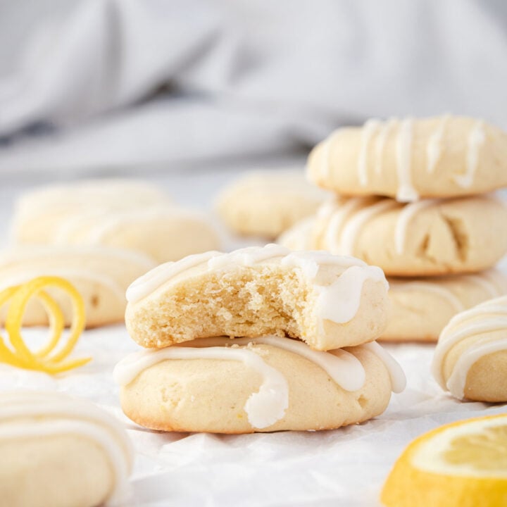 Two cookies stacked on each other with 1 bite out of the top one, curled lemon peel, and stacks of cookies around.