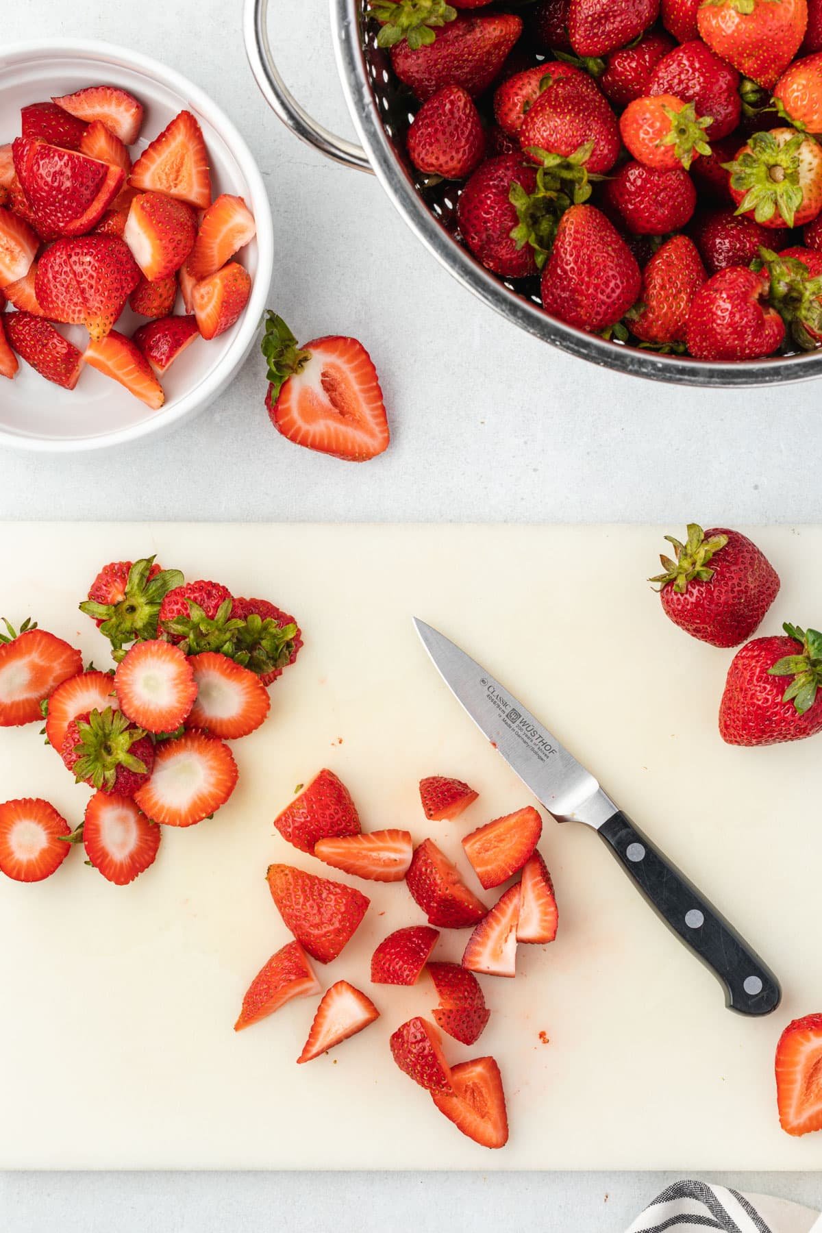 Curring board with a few whole strawberries on the right, sliced strawberries with a pairing knife in the middle, and the cut tops on the left.
