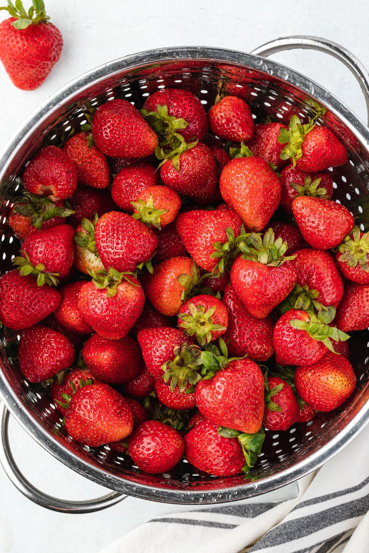 A large colander filled with freshly rinsed strawberries and one berry on the counter.