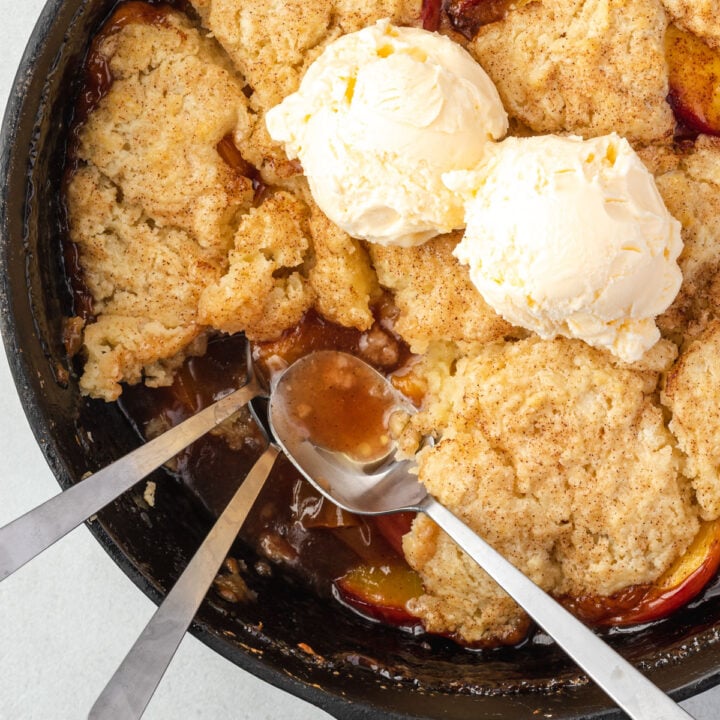 Close up of peach cobbler with 2 scoops of ice cream and 3 spoons digging in.