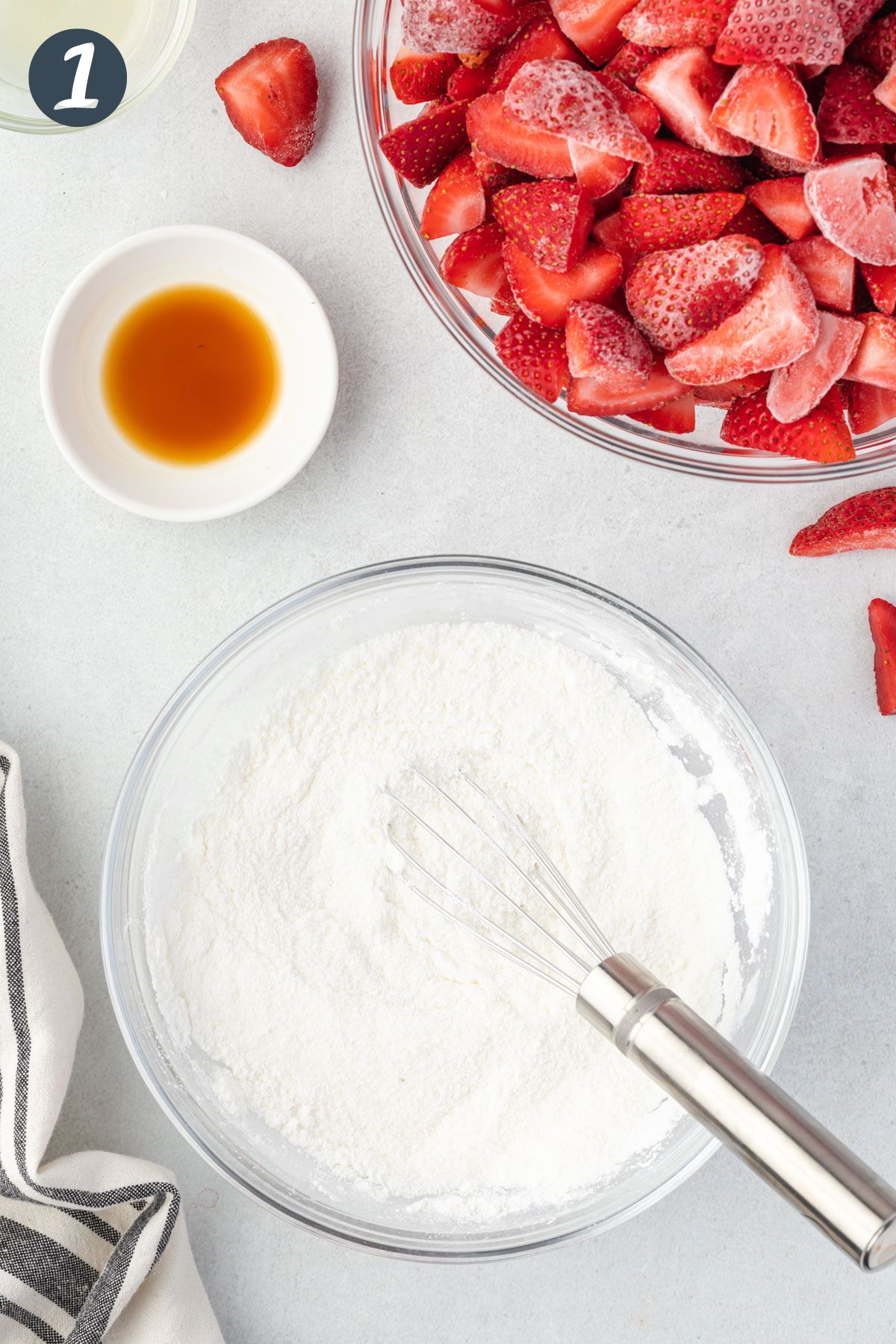 Sugar and cornstarch in a bowl with a whisk. Vanilla extract and strawberries in bowls at top.