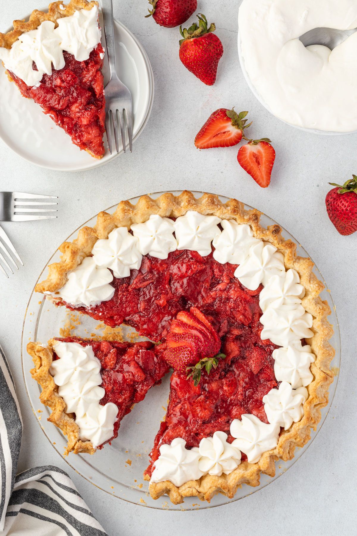Pie with a slice missing and a slice of pie on a plate with strawberries scattered around.