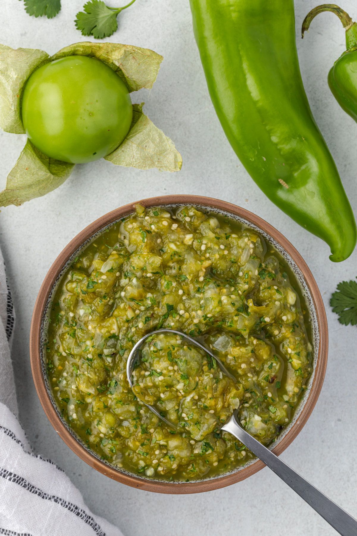 Overhead of a bowl of hatch salsa with a spoon, tomatillo and hatch pepper on top.