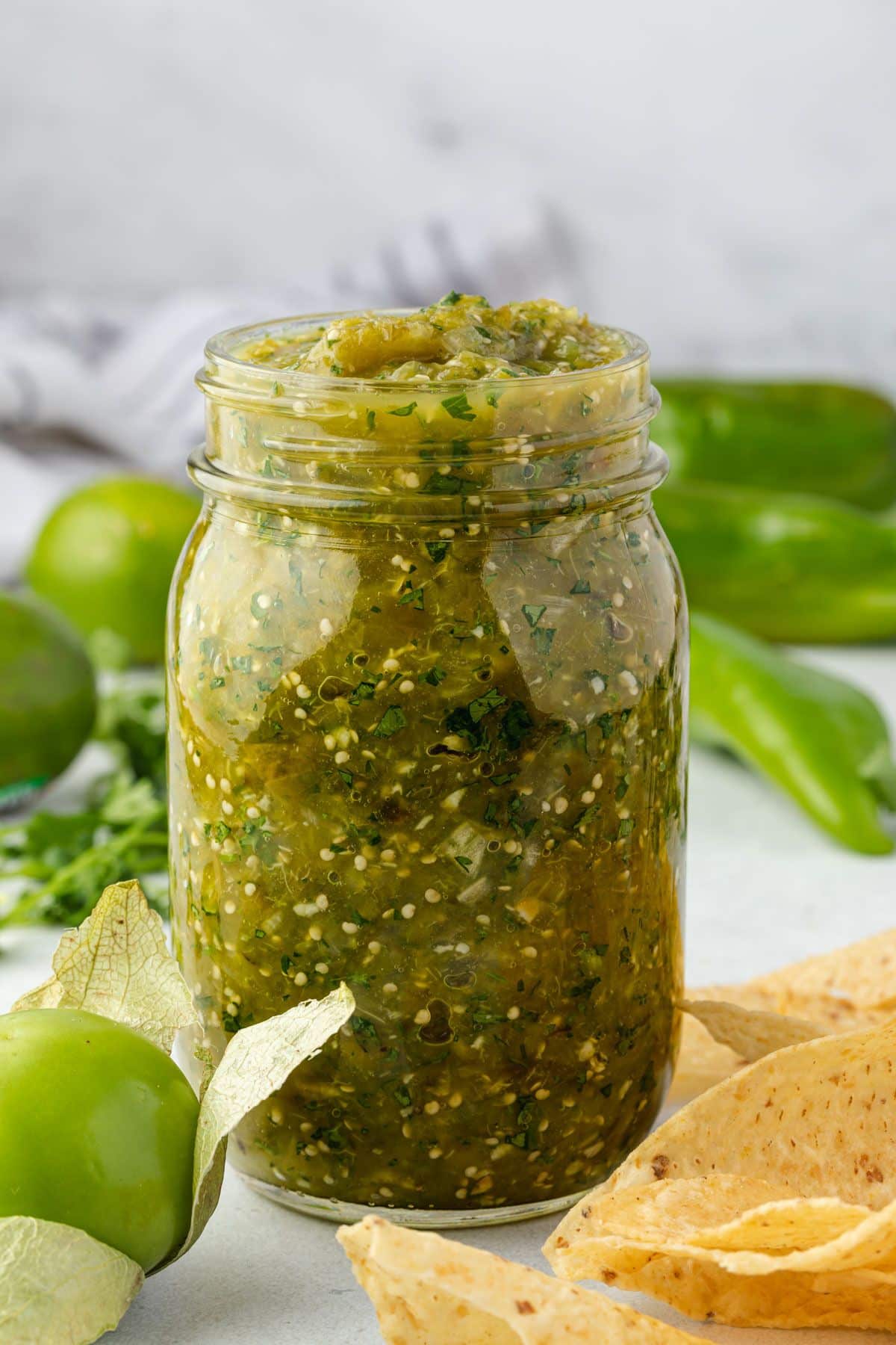 Jar of salsa verde with salsa ingredients and chips surrounding the jar.