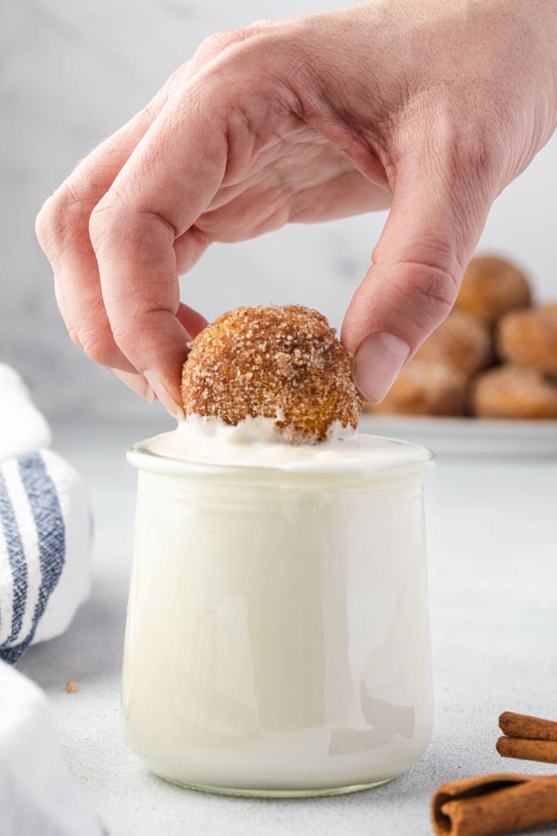 A hand holding a donut hole and dipping it into a jar of sweet cream cheese sauce.